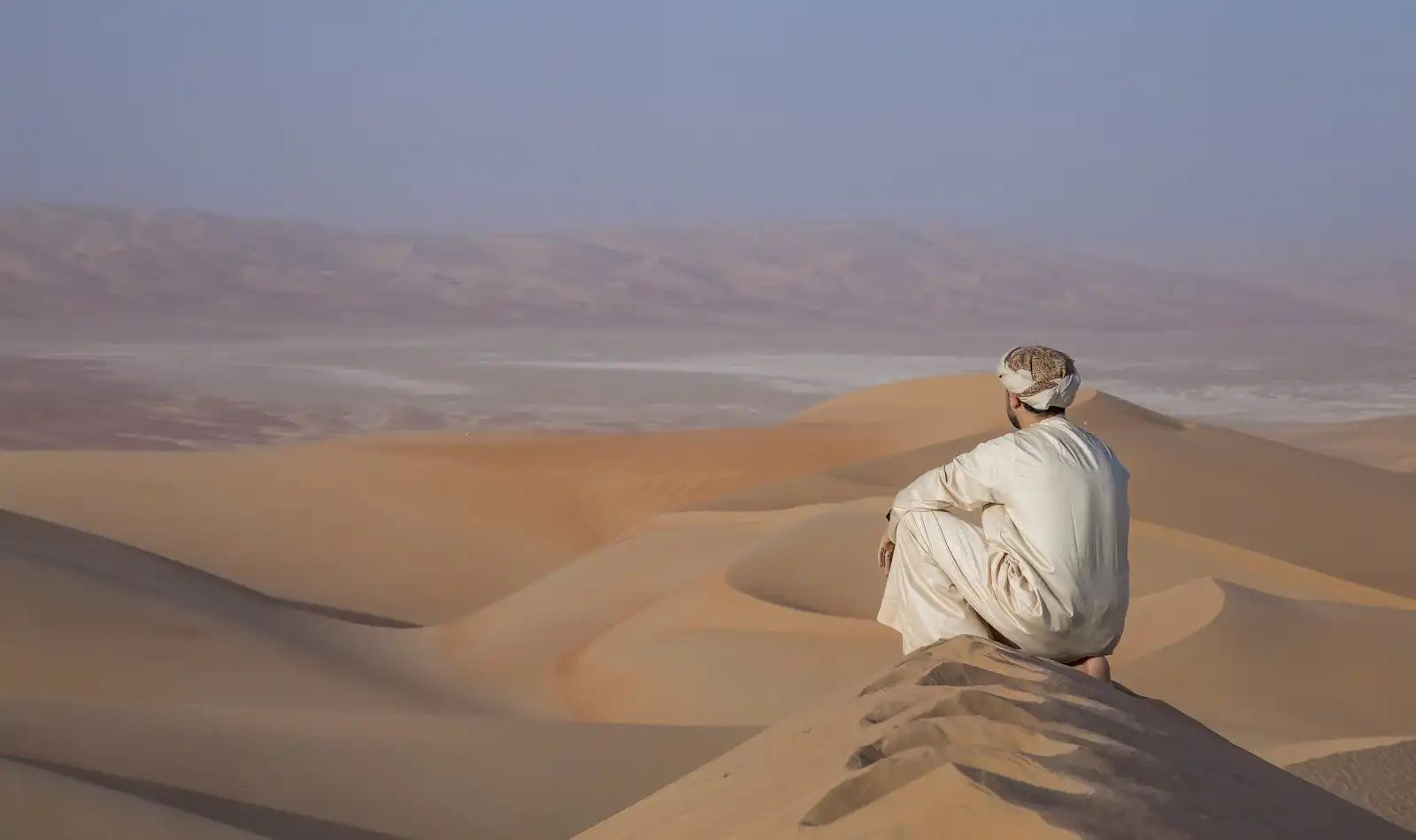 Man in traditional clothing sitting on a sand dune.